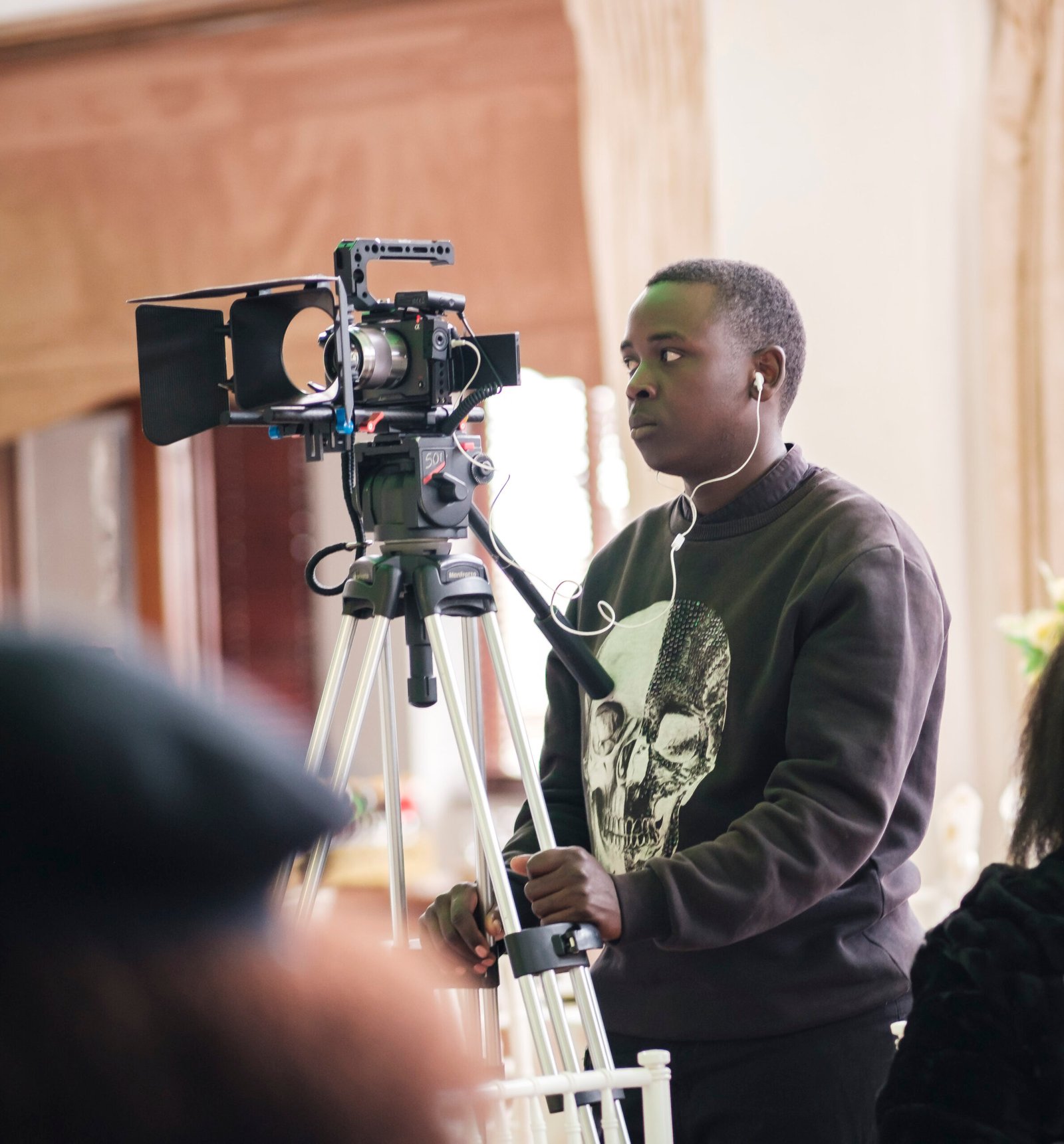 A person wearing headphones operates a professional video camera on a tripod indoors, focusing intently on the shot.